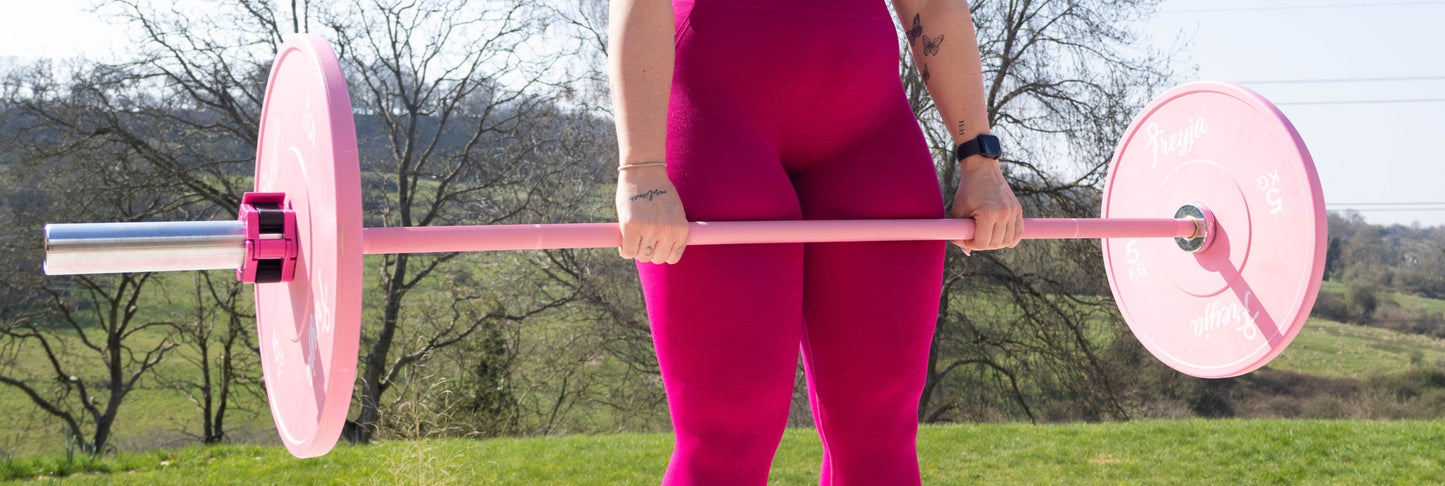Woman holding a pink freyja barbell  and bumper plates outdoors with trees and grass in the background