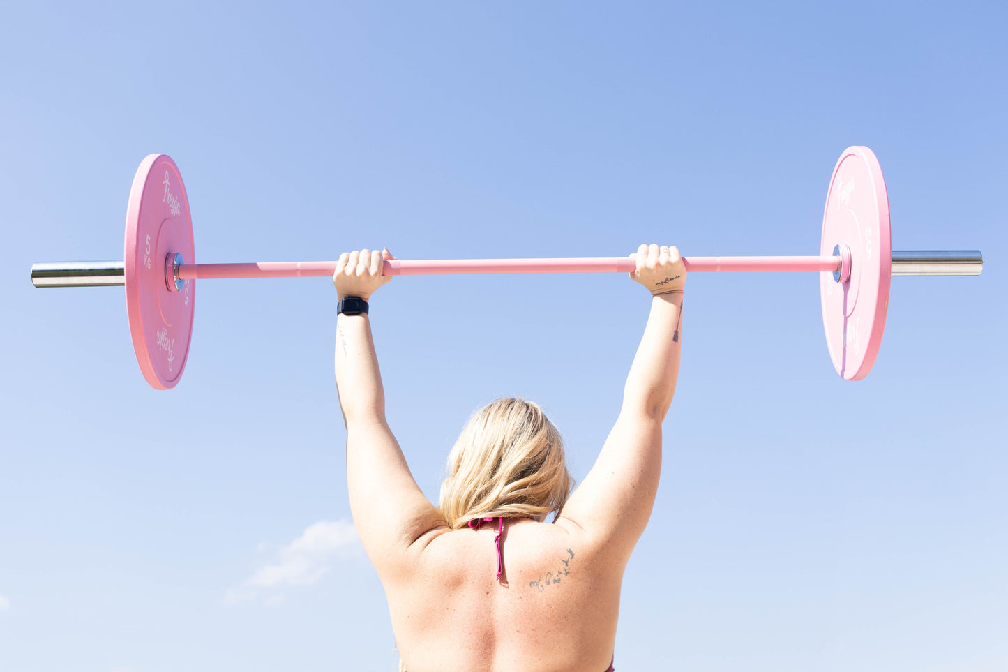 Woman lifting a pink Freyja Fit barbell loaded with bumper plate weights against a clear blue sky