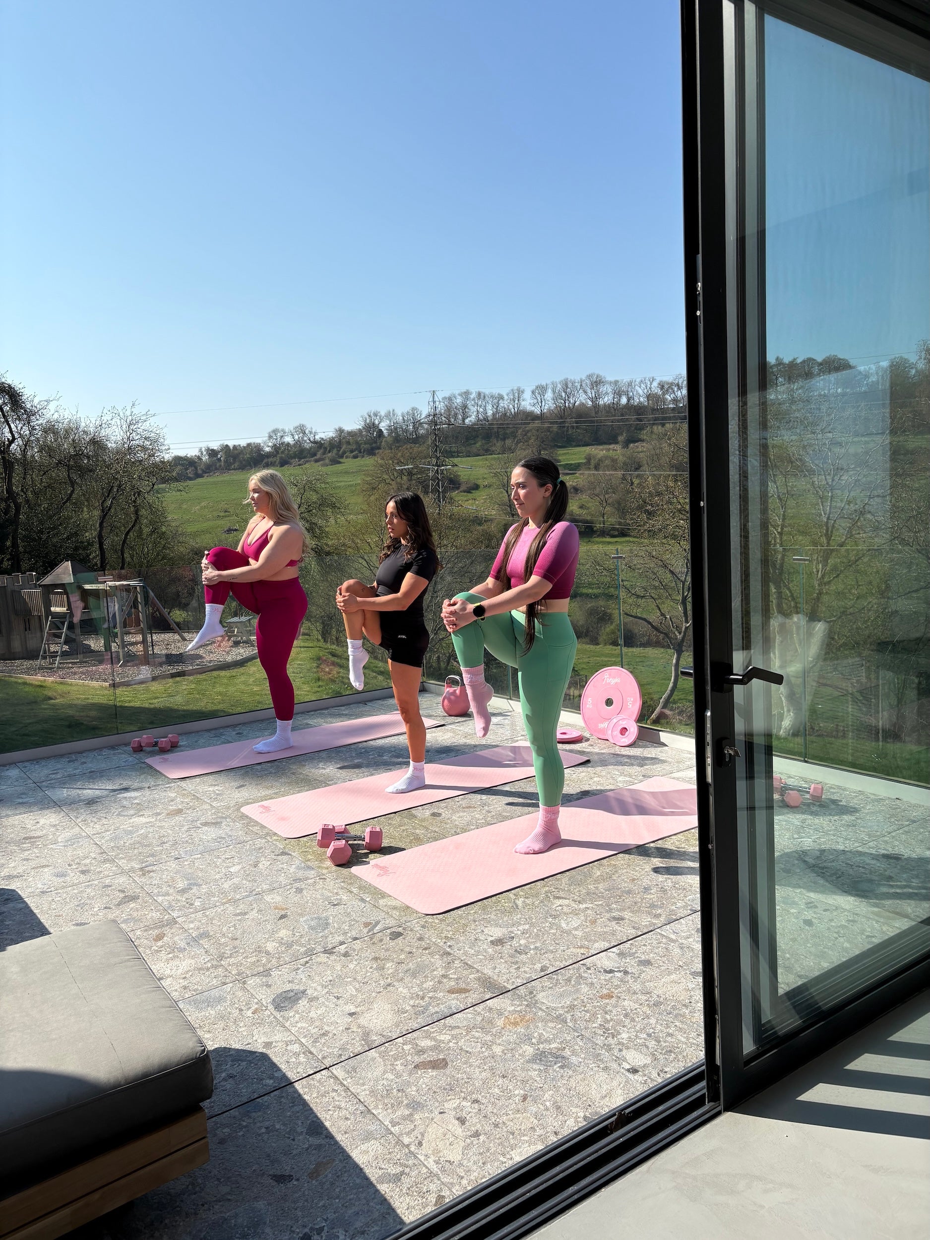 Three women exercising on pink Freyja  yoga mats outdoors with a scenic background.