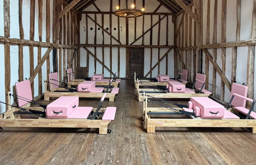A large rustic barn or studio with exposed wooden beams and a wood floor, showing multiple pink Pilates reformer machines arranged in two rows.
