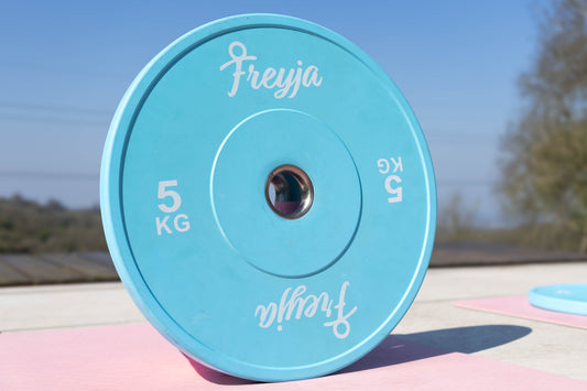 Blue weight plate with 'Freyja' branding on a pink yoga mat and a blurred outdoor background