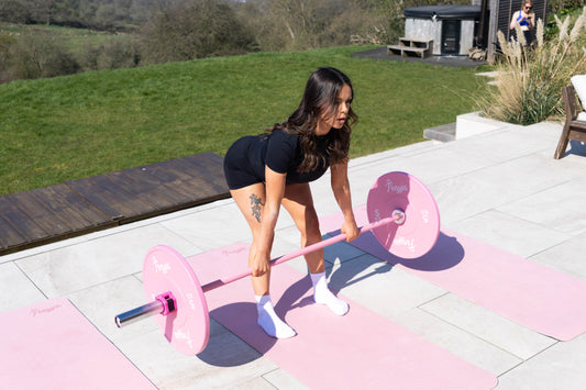 Woman lifting a pink Freyja Fit barbell with weighted plates outdoors on a pink yoga mat on a sunny day