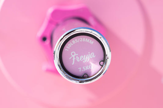 Close-up of a Freyja Fit barbell with branding visible on the end, with a blurred adjustable collar and weight plate in the background