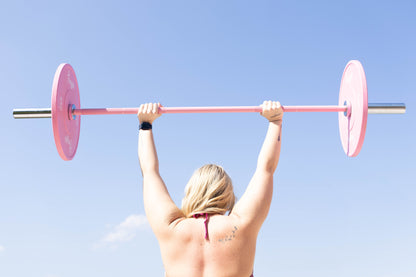Woman lifting a pink Freyja Fit barbell loaded with bumper plate weights against a clear blue sky