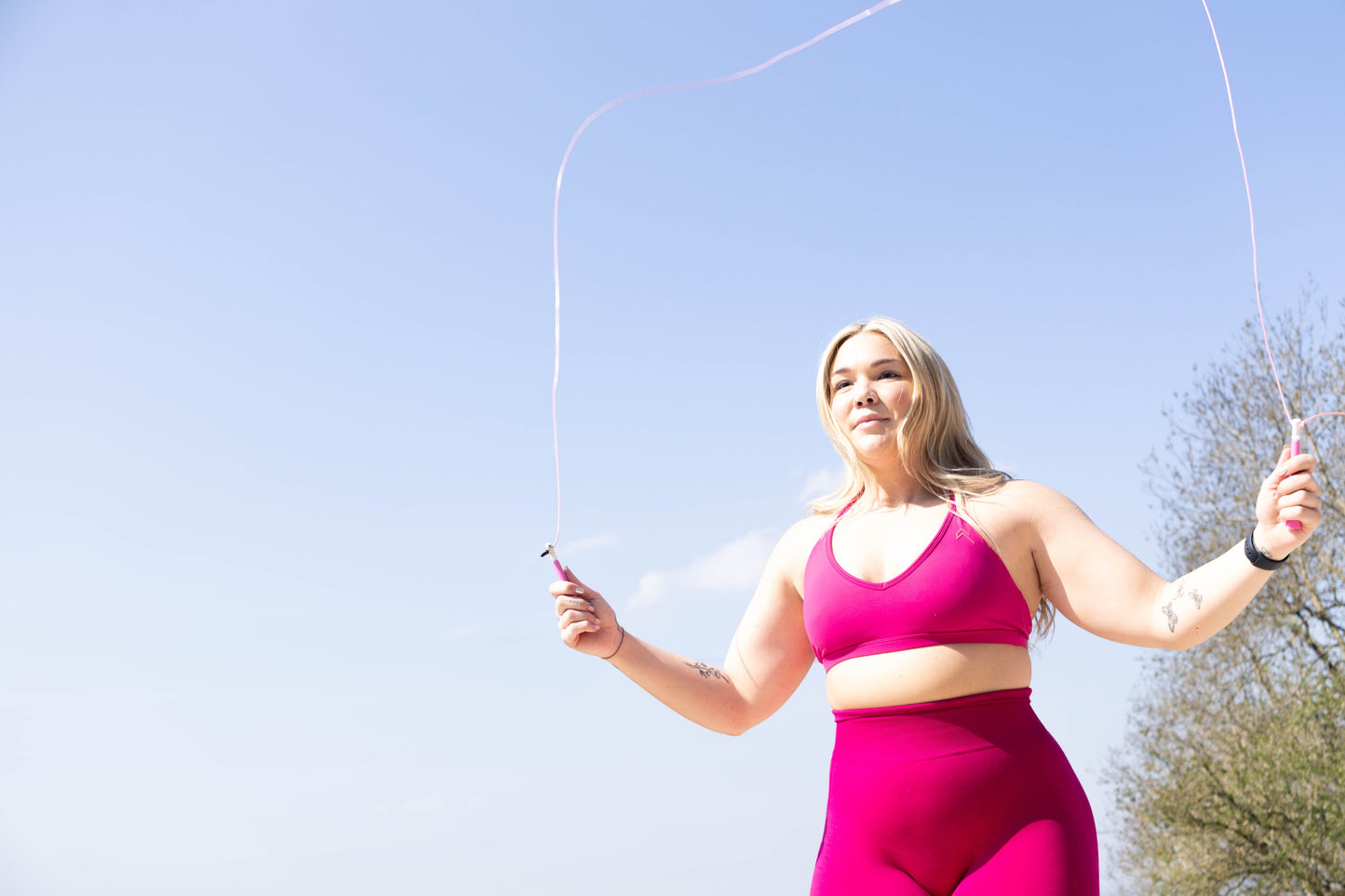 Woman in pink athletic wear using a pink Freyja skipping rope outdoors with a clear blue sky.