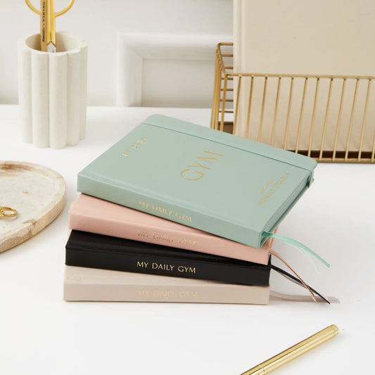 Stack of pastel-colored daily gym planners with gold lettering on a white desk with office supplies nearby