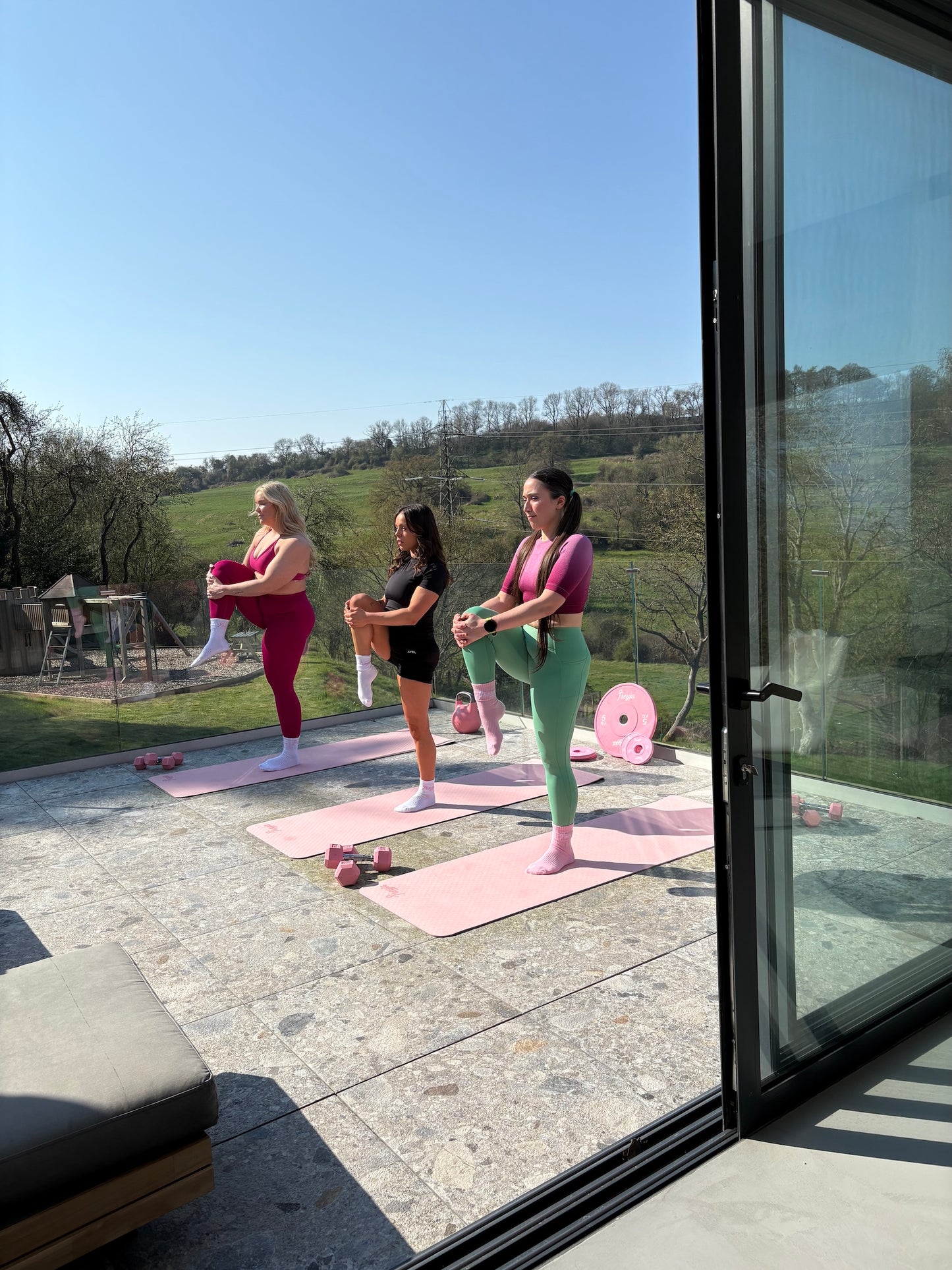 Three women exercising on pink Freyja  yoga mats outdoors with a scenic background.