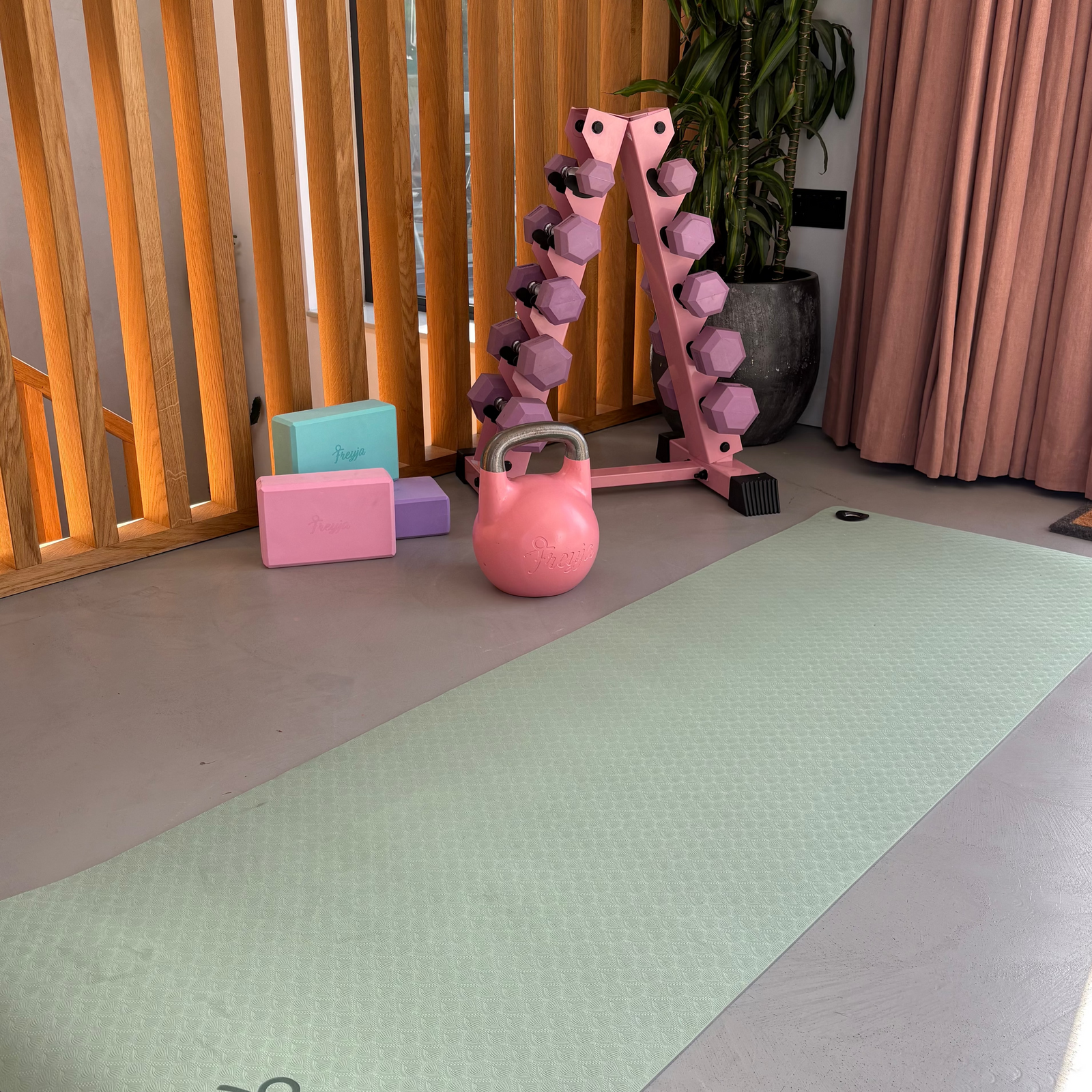 A pale mint green yoga mat with the name 'Freyja' printed in black script near one end, laid out on a gray floor in a home gym setting with a pink dumbbell rack, kettlebell, and yoga blocks.
