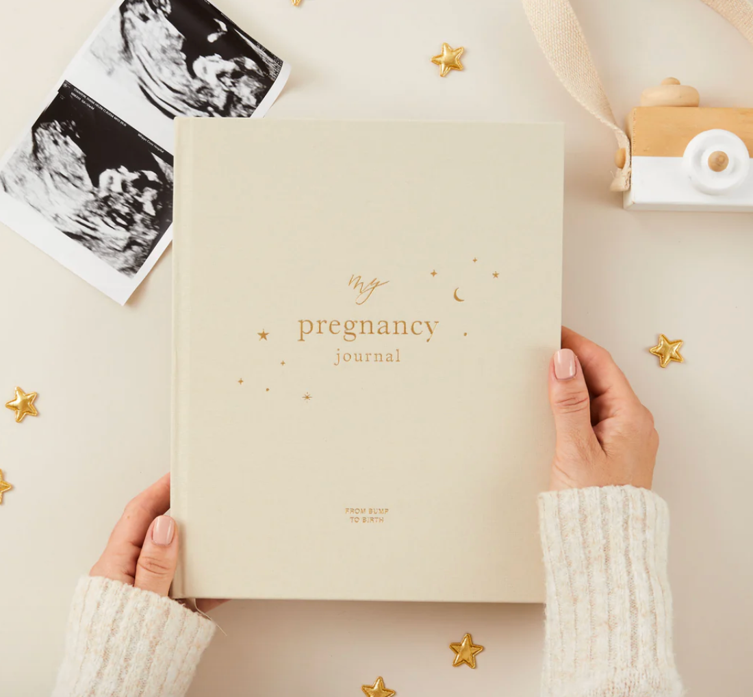 Hands holding a beige pregnancy journal surrounded by baby ultrasound photos and small gold star decorations on a table