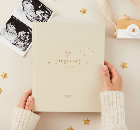 Hands holding a beige pregnancy journal surrounded by baby ultrasound photos and small gold star decorations on a table
