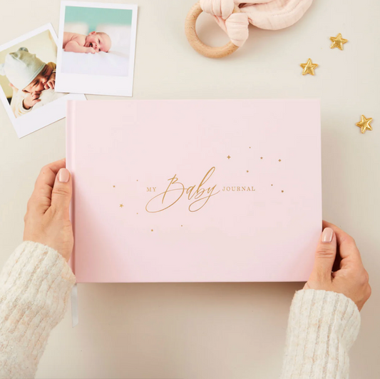 Hands holding a pink My Baby Journal book surrounded by baby photos, toy rattle, and gold star decorations