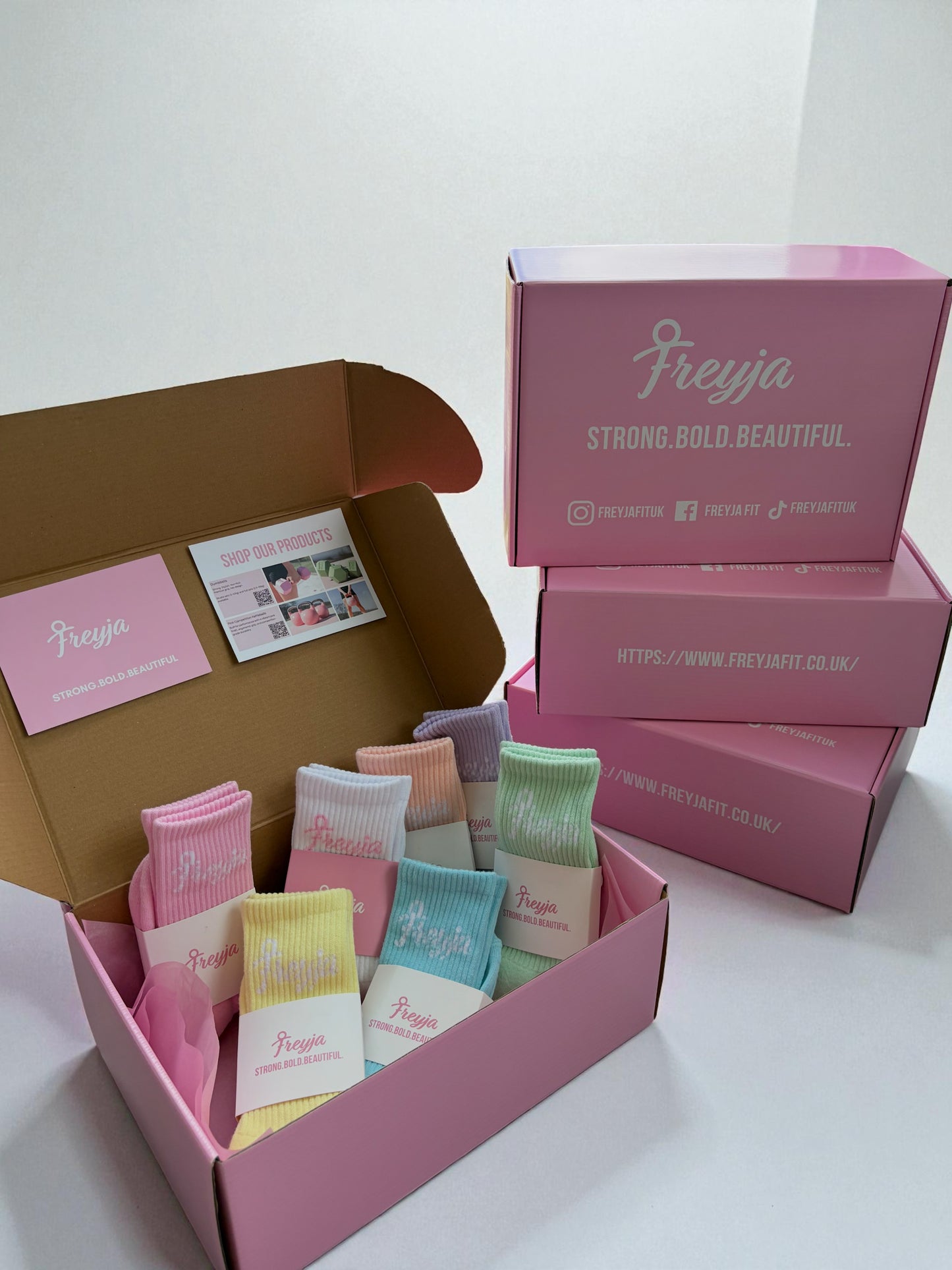 Open pink box displaying seven pairs of pastel-colored crew socks neatly arranged with branded packaging cards.