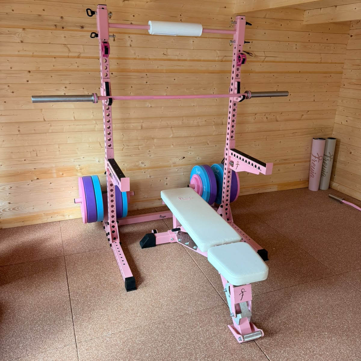 Pink home gym equipment set including a pink power rack with a barbell, weight plates, and a pink adjustable weight bench with a white padded surface, placed in a wooden room with wood plank walls and floor.